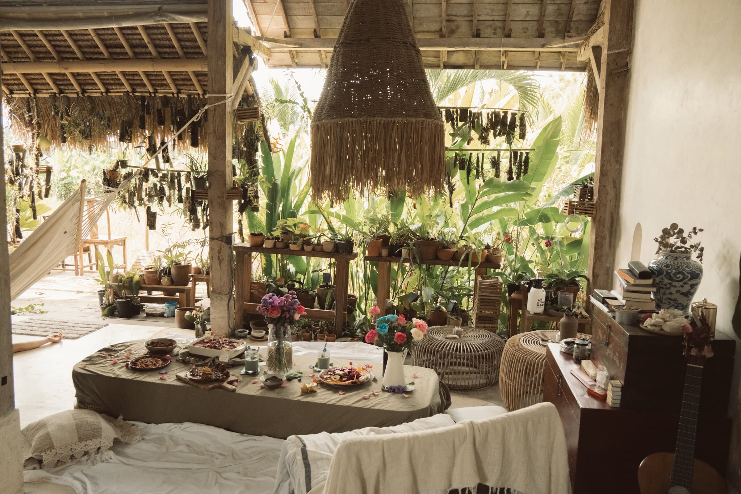 A communal bohemian gathering — long table set with flowers, plates, and candles inside an open pavilion surrounded by plants.