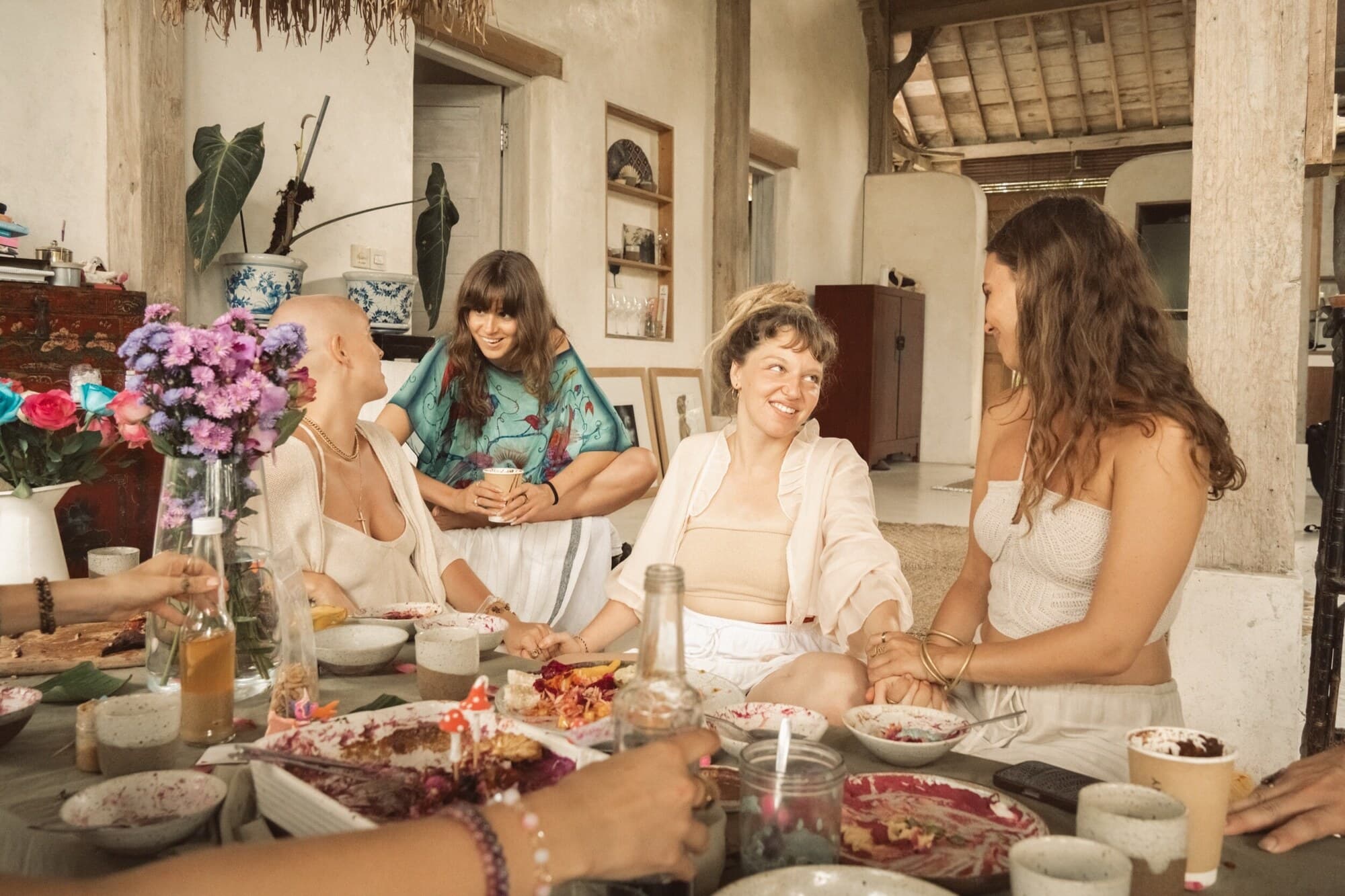 Four tribe members sitting around a low table of shared bowls and flowers at a Fairy Gathering in Bali, holding hands and laughing together.