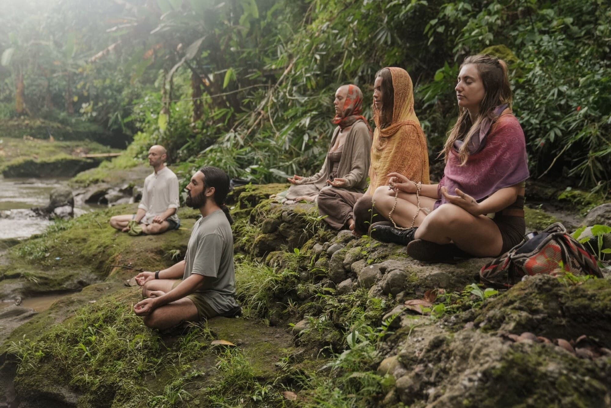 Tribe members sitting in meditation on mossy rocks beside a jungle stream.